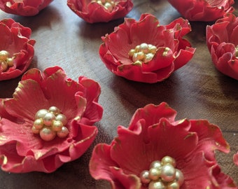 May include: Red sugar flowers with gold accents and pearl-like decorations. The flowers are arranged on a wooden surface.