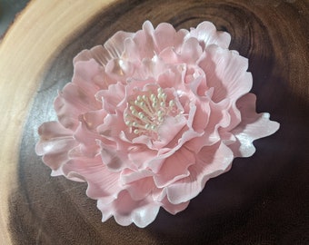 May include: A delicate pink peony flower with a white center, made of fondant. The flower is on a wooden surface.