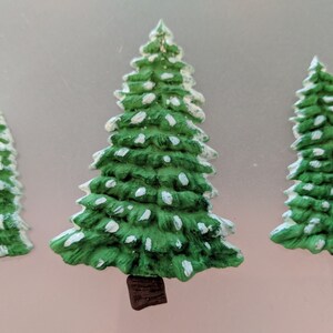 May include: Three green ceramic Christmas trees with white snow accents. The trees are different sizes and have brown bases.