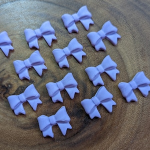 May include: A collection of 12 purple bow-shaped cake decorations. The bows are made of fondant or gum paste and are perfect for adding a touch of elegance to cakes, cupcakes, and other desserts.