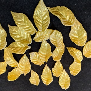 May include: A collection of gold-colored artificial leaves with detailed veining. The leaves vary in size and shape, and are arranged on a dark surface. The leaves have a metallic sheen, suggesting they are made of a shiny material.