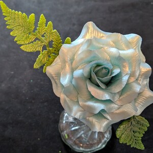 May include: A light blue silk rose with a green fern stem in a clear glass vase.