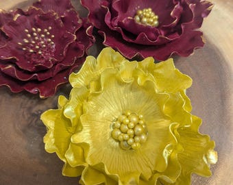 May include: Three decorative flower ornaments. Two are a deep burgundy color with gold accents, and one is a bright yellow. Each flower has a textured, layered petal design and a cluster of small, round beads in the center.