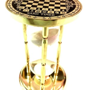 Puede incluir: Un reloj de arena de latón con una placa superior circular que presenta un patrón de tablero de ajedrez en blanco y negro. La placa superior tiene grabada la inscripción THE CHESS MAKER 6th CENTURY AD y SAND TIMER. Tres pilares verticales.