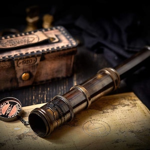 May include: A brass nautical telescope with a matching rectangular storage case. The telescope features a detached circular cap engraved with "DOLLOND LONDON 1920". The items are positioned on a vintage-style map with visible grid lines and text.