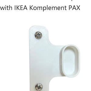 Può includere: Un supporto per asta appendiabiti bianco stampato in 3D con un alloggiamento ovale, dotato di due viti a croce color argento fissate sulla piastra di montaggio piatta. Il testo sopra recita: Clothes Rail Holder Compatible with IKEA Komplement PAX.