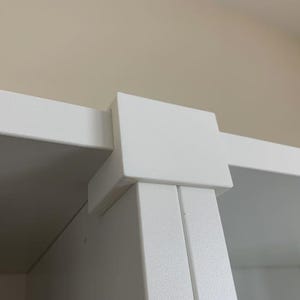 Peut inclure: Une pièce de raccordement de meuble en plastique blanc dotée d'un capuchon supérieur carré et d'un support latéral rectangulaire. Ce composant est conçu pour assembler deux panneaux blancs plats à angle droit, assurant une fixation solide.