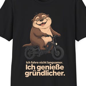 Puede incluir: Una camiseta negra de manga corta con un gráfico de una nutria marrón montando una bicicleta eléctrica negra. Debajo de la ilustración, el texto dice: Ich fahre nicht langsamer. Ich genieße gründlicher. El diseño está centrado en la parte frontal.