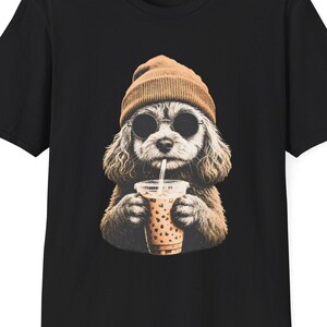 Op de afbeelding: Een zwart t-shirt met korte mouwen en een grafische print van een pluizige hond met een beige muts en een ronde zwarte zonnebril, die een doorzichtige plastic beker met een lichtbruine drank en een rietje in de voorpoten houdt.