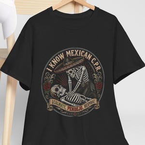 May include: A black t-shirt featuring a circular graphic of two skeletons performing CPR. The text reads I KNOW MEXICAN C.P.R. and ¡CALMATE, PENDEJO, RESPIRA!. The design includes a sombrero, red roses, and decorative scrollwork elements.