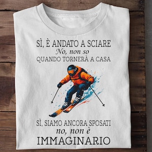 Könnte beinhalten: Ein weißes T-Shirt mit einer Grafik eines Skifahrers in orangefarbener Jacke und dunkler Hose. Der Text lautet: SÌ, È ANDATO A SCIARE No, non so QUANDO TORNERÀ A CASA SÌ, SIAMO ANCORA SPOSATI no, non è IMMAGINARIO. Das Design enthält blaue und orange Akzente.