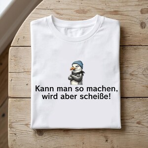 May include: A white t-shirt featuring a graphic of a seagull wearing a blue beanie and a scarf with its arms crossed. Below the graphic, the text reads: Kann man so machen, wird aber scheiße!