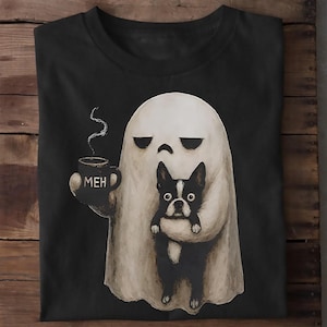 May include: A black t-shirt featuring a graphic of a white ghost holding a steaming mug with the text MEH printed on it. The ghost is also holding a black and white Boston Terrier dog. The design is centered on the front of the garment.