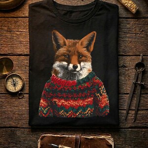Puede incluir: Una camiseta negra con una ilustración gráfica de un zorro que lleva un suéter de punto multicolor con patrones rojos, verdes y naranjas. El zorro tiene pelaje marrón, marcas blancas en el pecho y mira directamente hacia adelante.