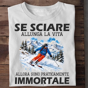 May include: A white t-shirt featuring a graphic of a skier on a snowy mountain slope. The text above reads SE SCIARE ALLUNGA LA VITA and the text below reads ALLORA SONO PRATICAMENTE IMMORTALE. The skier wears a blue jacket, orange pants, and a helmet.