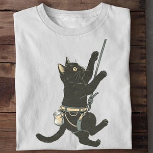 Puede incluir: Una camiseta blanca con una ilustración gráfica de un gato negro que lleva un arnés de escalada y una bolsa de magnesio, trepando por una cuerda. El gato aparece de perfil, estirando sus patas delanteras para agarrar la cuerda vertical.