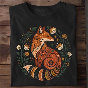 Könnte beinhalten: Ein schwarzes Kurzarm-T-Shirt mit einer zentralen Grafik eines Fuchses, der mit komplexen orangefarbenen, braunen und cremefarbenen Mustern verziert ist. Der Fuchs ist von einer kreisförmigen Anordnung stilisierter grüner Blätter und Blumen umgeben.