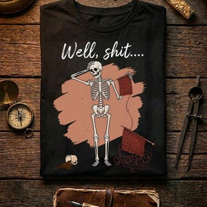 May include: A black t-shirt featuring a graphic of a white skeleton holding knitting needles and a piece of red knitted fabric. Above the skeleton, the text reads "Well, shit...." A spilled mug of brown liquid is visible near the skeleton's feet.