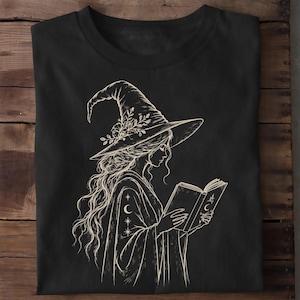 Könnte beinhalten: Ein schwarzes T-Shirt mit einer cremefarbenen Strichzeichnung einer Person, die einen mit Blumen verzierten Hexenhut mit breiter Krempe trägt. Die Figur hat langes, welliges Haar und hält ein offenes Buch mit Mond- und Sternsymbolen in den Händen.