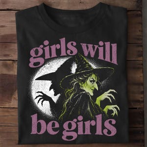 Può includere: Una t-shirt nera con la grafica di una strega dalla pelle verde con un cappello a punta davanti a una grande luna bianca. Il testo "girls will be girls" è stampato in un carattere viola consumato sopra e sotto l'illustrazione della strega.