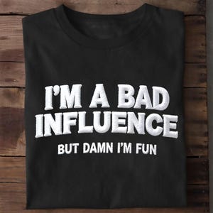 May include: A black short-sleeved t-shirt featuring white embroidered text that reads I'M A BAD INFLUENCE BUT DAMN I'M FUN. The lettering is arranged in three lines with the first two lines in a large, bold, sans-serif font style.