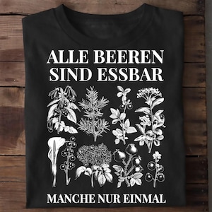 Könnte beinhalten: Ein schwarzes T-Shirt mit kurzen Ärmeln und weißen botanischen Illustrationen verschiedener Beeren und Pflanzen. Der Text "ALLE BEEREN SIND ESSBAR" steht oben und "MANCHE NUR EINMAL" unten in einer klaren, weißen Schriftart.