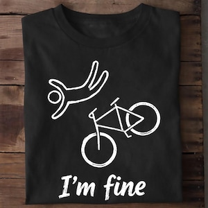 Op de afbeelding: Een zwart T-shirt met korte mouwen en een witte afbeelding van een stokfiguur die door de lucht valt boven een fiets. Onder de afbeelding staat de tekst "I'm fine" gedrukt in een wit, licht verweerd schreefloos lettertype.
