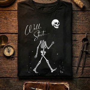 May include: A black t-shirt featuring a white graphic of a headless skeleton walking while holding a skull on a string like a balloon. The text "Well. Shit...." is printed in a white cursive script above the skeleton figure on the front.