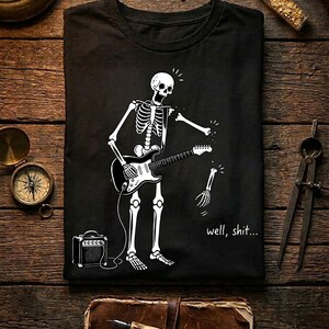 May include: A black t-shirt featuring a white graphic of a skeleton playing an electric guitar. The skeleton's left arm is detaching at the shoulder, with the hand and forearm falling away. The text "well, shit..." is printed near the bottom right.