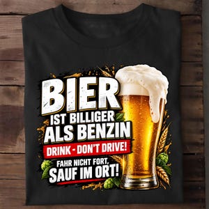 Könnte beinhalten: Ein schwarzes T-Shirt mit einer Grafik eines goldenen Bierglases mit weißem Schaum. Der Text lautet: BIER IST BILLIGER ALS BENZIN, DRINK - DON'T DRIVE!, FAHR NICHT FORT, SAUF IM ORT! Das Design zeigt zudem Abbildungen von Weizenähren und grünen Hopfendolden.