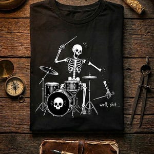 May include: A black t-shirt featuring a white graphic of a skeleton playing a drum set. The skeleton is missing an arm, with the detached limb holding a drumstick. The text "well, shit..." is printed in white near the bottom right of the graphic.