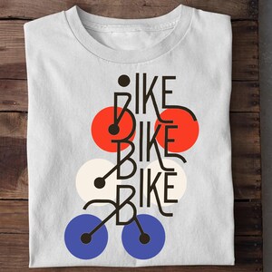 May include: A white short-sleeved t-shirt featuring a graphic design with the word BIKE repeated three times in black lettering. The design incorporates stylized bicycle shapes and three pairs of colored circles in red, off-white, and blue.