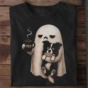Könnte beinhalten: Ein schwarzes T-Shirt mit einer Grafik eines weißen Geistes, der eine schwarze Tasse mit der Aufschrift MEH hält. Der Geist hält zudem einen kleinen schwarz-weißen Hund mit großen Augen. Die Illustration hat eine handgezeichnete, texturierte Optik.