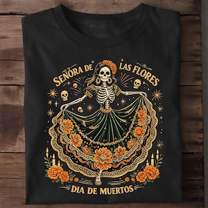 May include: A black t-shirt featuring a graphic of a skeleton wearing a long, dark green dress adorned with orange flowers. The text reads "SEÑORA DE LAS FLORES" and "DIA DE MUERTOS" in yellow, surrounded by small skulls, candles, and starbursts.