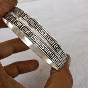 May include: Two silver-toned metal bangles featuring a continuous embossed Greek key pattern. The bracelets have a polished, reflective finish on the inner bands and a darkened, recessed design on the outer surface, creating a high-contrast look.