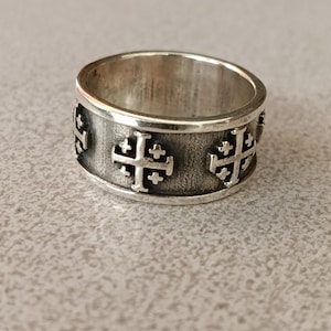 Puede incluir: Un anillo de banda ancha de plata con un patrón repetitivo de cruces de Jerusalén en relieve. El metal tiene un acabado pulido en los bordes y un fondo texturizado y oscurecido en las áreas hundidas para resaltar los símbolos.