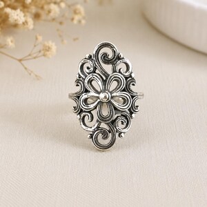 May include: A silver-toned ring featuring an elongated oval face with an intricate openwork design. The center displays a six-petaled flower motif surrounded by swirling filigree patterns, with darkened recessed areas to highlight the metal details.