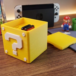 Peut inclure: Un conteneur de rangement jaune en forme de cube avec un point d'interrogation blanc sur la face avant, conçu pour contenir des cartouches de jeux vidéo. Le récipient possède un couvercle amovible et une finition texturée avec des fentes internes.