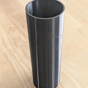 May include: A tall, cylindrical, dark gray vessel with a smooth, uniform exterior and a hollow interior. The object features vertical striations along its outer surface, indicating a layered construction process, and has a simple, open circular rim.