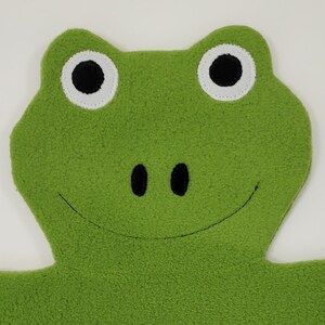 May include: A plush green frog face with large white-rimmed eyes and a smiling mouth. The frog has two black oval nostrils. The soft, textured material suggests a children's toy or decorative item.