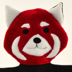May include: A plush red panda head with white accents around the eyes and mouth, and white ears with black tips. The panda has black eyes and a black nose. The bottom portion of the panda is black.