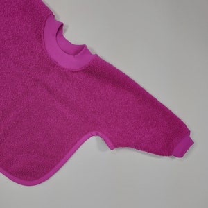 May include: A pink terry cloth bib with a round neckline and a single long sleeve.