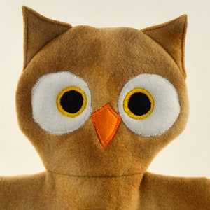 May include: A brown owl-shaped plush toy with large white eyes, black pupils, and yellow accents. The owl has an orange beak and pointed ears. The toy is made of soft, felt-like material and is likely handmade.