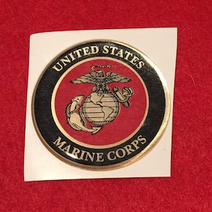 May include: A circular sticker featuring the United States Marine Corps emblem. The design includes a globe, eagle, and anchor, surrounded by the words "United States Marine Corps" in a black and gold border on a red background.