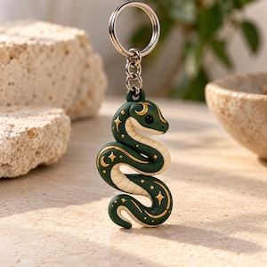 Pode incluir: Um porta-chaves de cobra verde-escura com uma barriga cor de creme e motivos celestiais dourados, incluindo estrelas e luas crescentes. A cobra está presa a um anel de metal prateado por um conector de corrente curto.