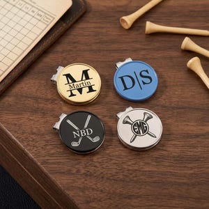 May include: Four round metal golf ball markers with clips. One gold marker features the name Martin, one blue marker shows the initials DS, one black marker displays NBD with crossed golf clubs, and one silver marker shows EMG with crossed golf tees.