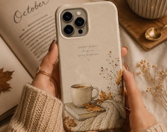 May include: A beige smartphone case featuring a printed illustration of a coffee mug resting on a book, surrounded by autumn leaves and a knit texture. The text reads: golden leaves, crisp air, quiet moments, with a small heart symbol below.