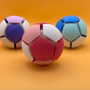 May include: Three spherical puzzle balls with a geometric pentagonal pattern. The front ball features pink and white segments. The left ball has purple and lavender segments. The right ball displays blue and light teal segments on a white base.
