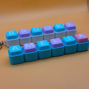 May include: Two custom keychains featuring stacked square blocks in alternating light blue and light purple colours. The top keychain spells BROOKE with white letters, and the bottom keychain spells AMELIA with white letters on the square blocks.