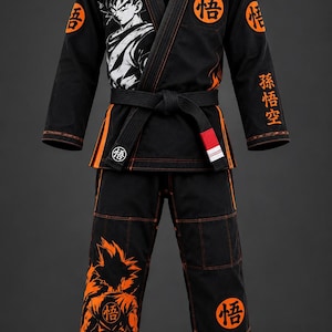 Op de afbeelding: Een zwart vechtsport-gi met oranje stiksels, voorzien van een afbeelding van een personage met stekelig haar op de borst en het been. Het kledingstuk bevat ronde oranje patches met zwarte Kanji-symbolen en verticale oranje Kanji-tekst op de mouw.